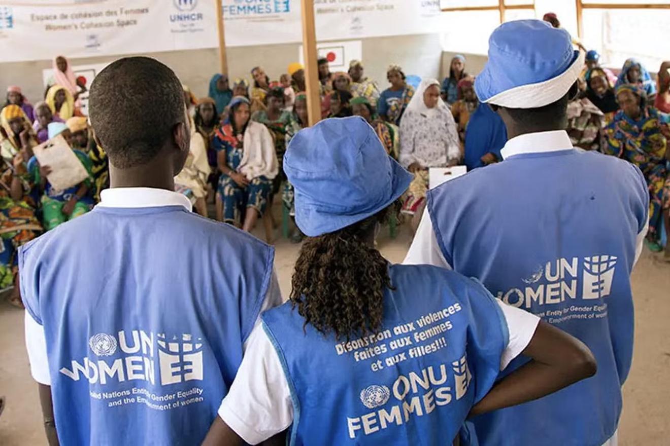 A group of people gathered in a community meeting. Three individuals, wearing blue vests and hats with "UN WOMEN" written on them, are standing at the front addressing the seated audience. 